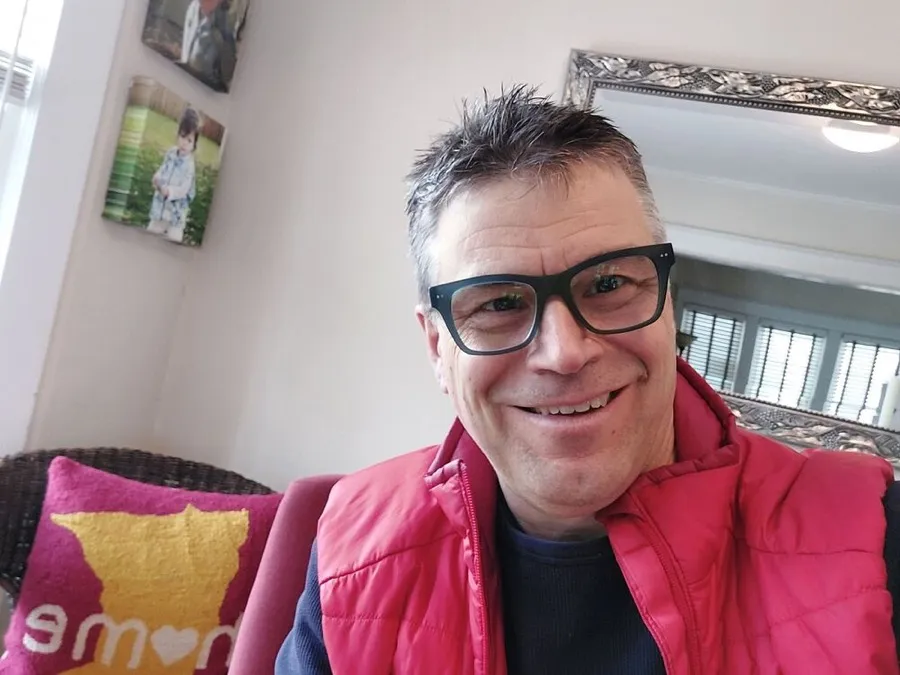 Michael smiling in LOOK OPTIC Cosmo readers, red vest, sitting on a couch