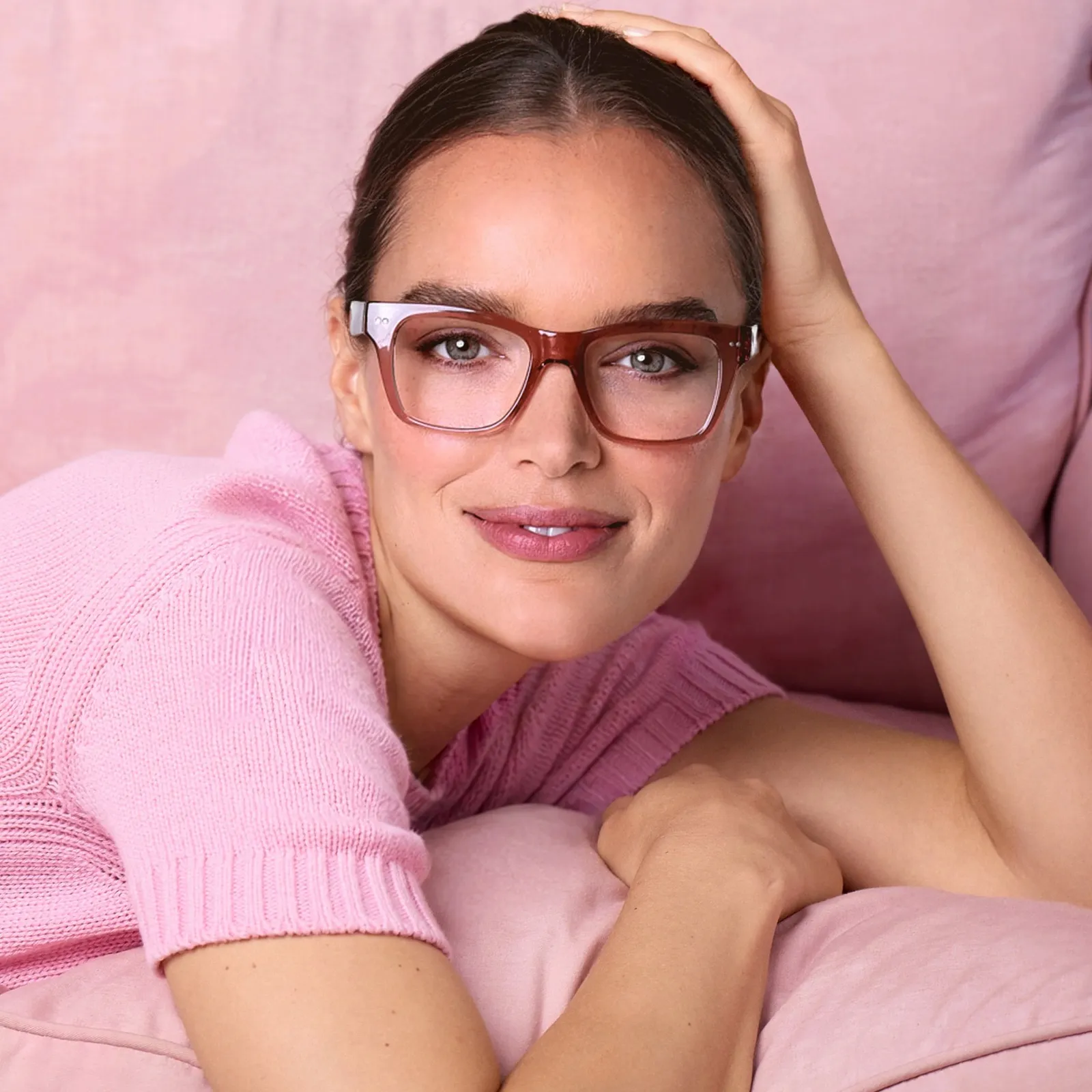 A smiling woman in a pink knit top, hand resting on her head as she lies on a pink sofa, wearing LOOK OPTIC Cosmo readers in Shiny Pink